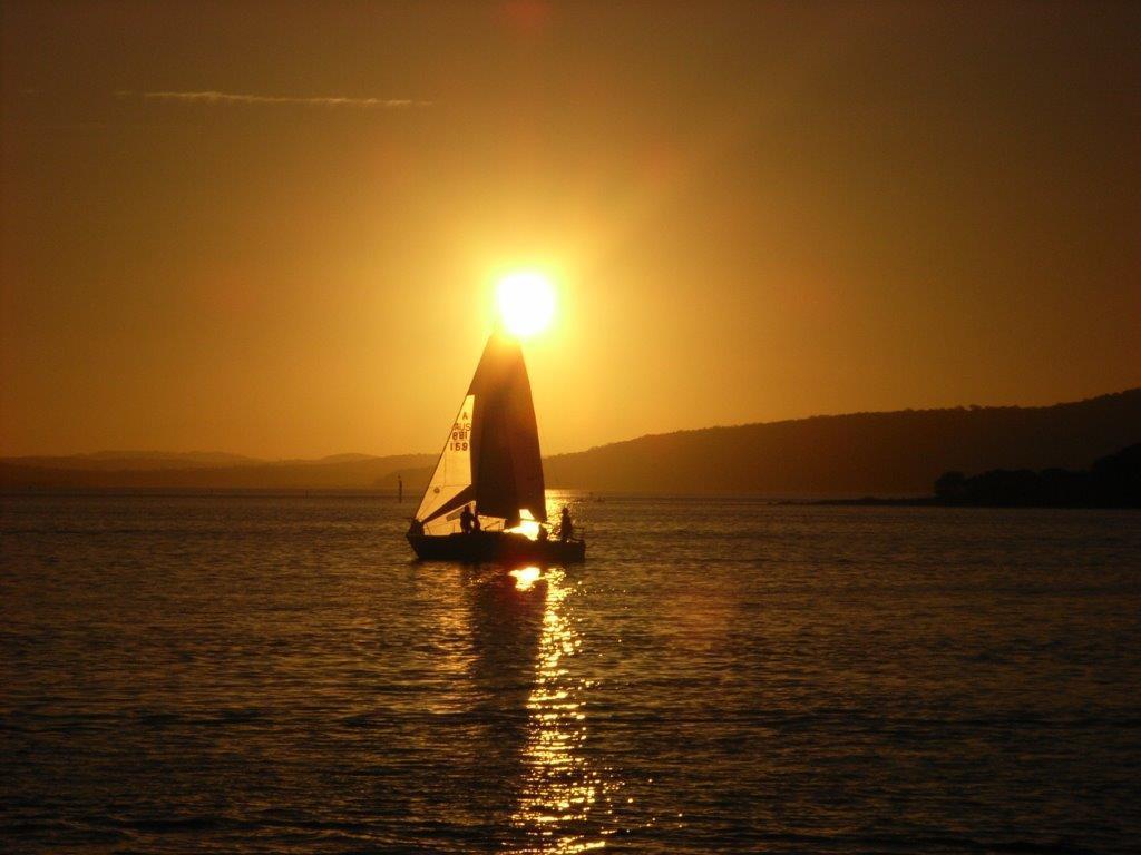 Silhouetted sailboat gliding across calm water during a golden sunset at Lake Macquarie